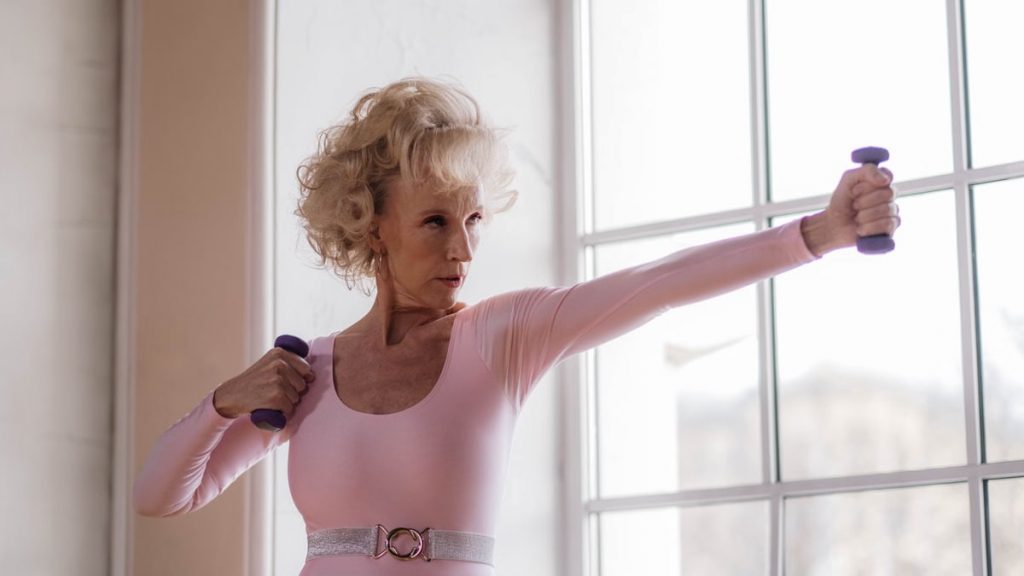 Senior woman in pink leotard working out with dumbbells near a large window indoors.